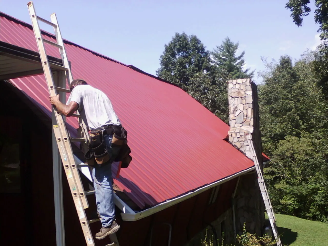 Roofer on a ladder beside a red metal roof and stone chimney — Soffit & Fascia Repair by Roofing Brothers in Monroe Township
